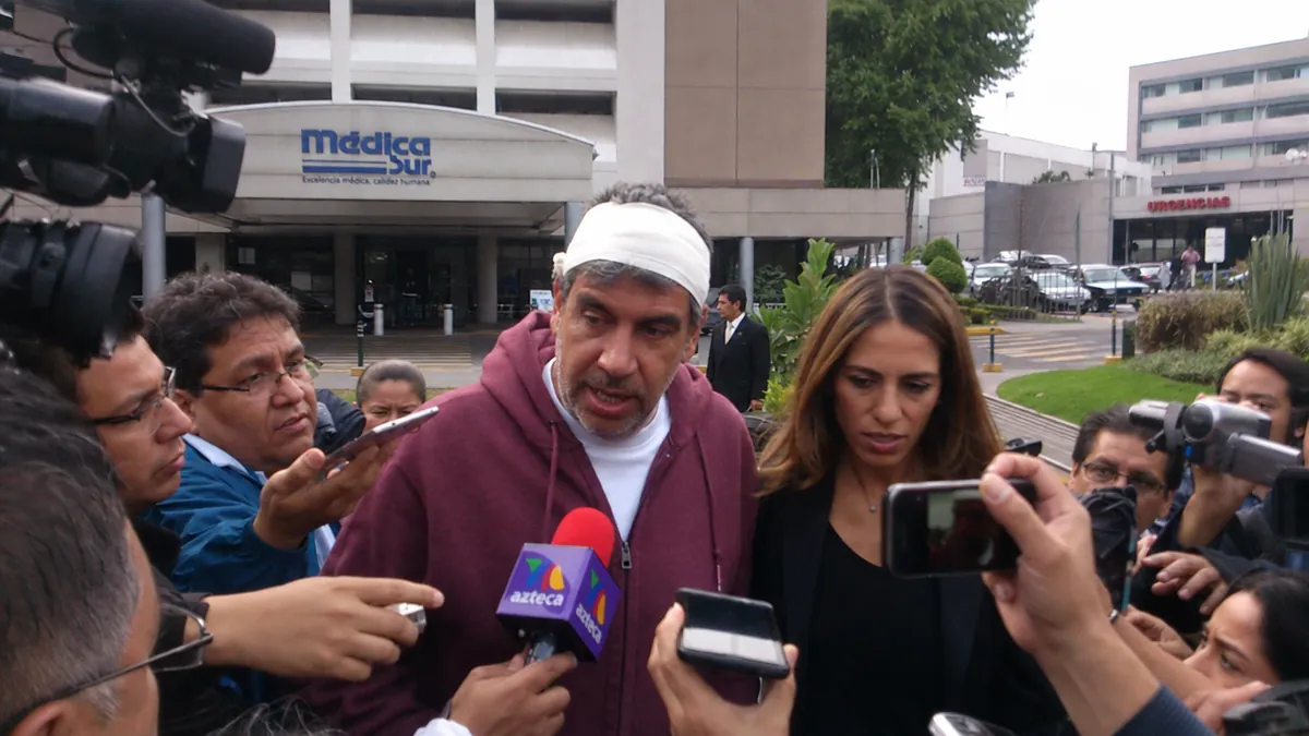 Arturo Elías Ayub hace declaración a la prensa a la salida del Hospital Médica Sur, con la cabeza vendada, rodeado de reporteros y micrófonos.